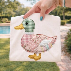 Peut inclure: Un t-shirt blanc avec un motif de canard en patchwork. Le canard a une tête à carreaux verts et blancs, un bec jaune et un corps composé de patchs de tissu à fleurs roses et rayées. Les pieds du canard sont jaunes.