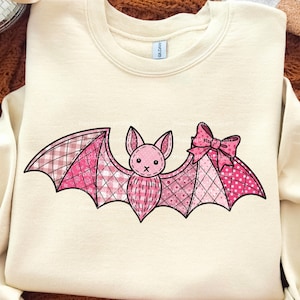 May include: Cream-colored sweatshirt featuring a pink bat design. The bat has patchwork wings with plaid and polka dot patterns, a pink bow, and a cute face. The design is centered on the front of the sweatshirt.
