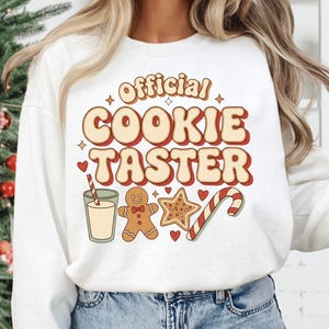 May include: White sweatshirt with the text "Official Cookie Taster" in a retro font. The design includes illustrations of a glass of milk with a straw, a gingerbread man, a star cookie, and a candy cane.