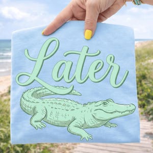May include: A light blue t-shirt featuring a green alligator illustration and the word "Later" in a cursive font. The design is centered on the shirt, with the alligator below the word. The shirt is held up by a person.