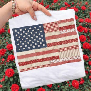 May include: White t-shirt featuring a patchwork American flag design. The flag incorporates various patterns, including gingham, floral, and striped fabrics, with a blue field of stars and red and cream stripes. The shirt is held up against a backdrop of red roses.