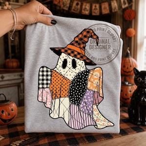 May include: A light gray t-shirt featuring a central graphic of a ghost wearing a witch hat. The ghost is designed with a patchwork pattern of various fabrics, including orange polka dots, black and white checks, and purple floral prints.