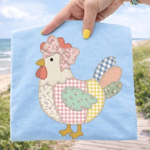 Könnte beinhalten: Hellblaues T-Shirt mit einem Patchwork-Hühnerdesign. Das Huhn hat eine rosa Blumenschleife, einen roten Kamm und einen Körper aus verschiedenen gemusterten Stoffen, darunter Vichy- und Blumendrucke. Der Hintergrund zeigt eine Strandszene.