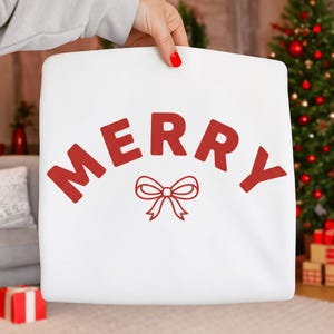 Könnte beinhalten: Weiße Tragetasche mit dem Wort "MERRY" in Rot, oben gewölbt, und einer roten Schleifengrafik darunter. Die Tasche wird von einer Person gehalten. Der Hintergrund umfasst einen Weihnachtsbaum und andere festliche Dekorationen.