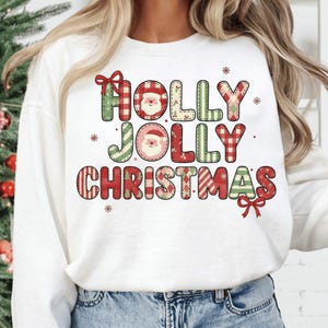 Peut inclure: Sweat-shirt blanc avec l'inscription "HOLLY JOLLY CHRISTMAS" dans un motif patchwork. Les lettres sont rouges, vertes et beiges avec des visages de Père Noël et des flocons de neige. Un nœud rouge est en bas à droite.