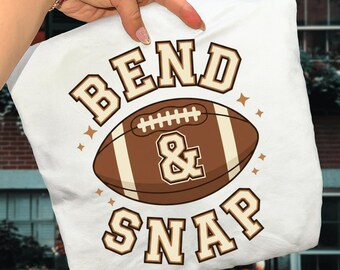 Op de afbeelding: Een wit t-shirt met een bruine American football-afbeelding in het midden. De tekst "BEND & SNAP" is in een lettertype in collegestijl boven en onder de football gedrukt. Kleine steraccenten omringen het centrale ontwerp.