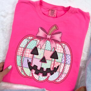 May include: A hot pink Comfort Colors sweatshirt featuring a patchwork pumpkin design. The pumpkin has a pink bow, a stitched outline, and a smiling jack-o'-lantern face. The pumpkin is made of pastel colored squares.
