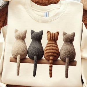 May include: Cream-colored sweatshirt featuring a graphic of four cats sitting on a wooden beam. The cats are in shades of beige, gray, and brown, with one displaying orange stripes. The cats are facing away from the viewer.
