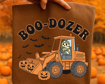 May include: Brown tote bag with the text "BOO-DOZER" in black and orange. The design features a cartoon ghost driving an orange bulldozer with pumpkins in the front and bats flying in the background. The background is an autumn scene.