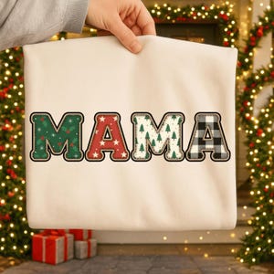 Könnte beinhalten: Ein cremefarbenes Sweatshirt mit dem Wort "MAMA" in großen, dekorativen Buchstaben. Jeder Buchstabe hat ein einzigartiges Muster: grünes Stechpalmenlaub, rote Sterne, grüne Weihnachtsbäume und schwarz-weißes Büffelkaro. Das Sweatshirt wird von einer Person gehalten.