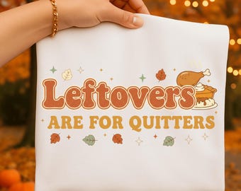 May include: White graphic tee with the text "Leftovers Are For Quitters" in orange and brown. The design includes illustrations of a turkey leg, pie, and fall leaves. The tee is held by a person with a gold bracelet.