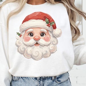 Op de afbeelding: Witte sweatshirt met een kleurrijke illustratie van de kerstman. De kerstman heeft een rode muts, roze wangen en een witte baard. Het ontwerp bevat hulstbladeren en bessen, perfect voor de feestdagen.