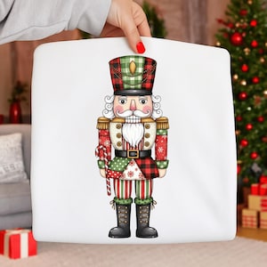 May include: A white square pillow featuring a colorful illustration of a traditional nutcracker soldier. The nutcracker wears a red, green, and black plaid hat and uniform, holding a candy cane. The design is festive and suitable for Christmas.
