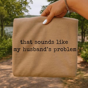 以下が含まれることがあります： 「that sounds like my husband's problem」という黒い文字がプリントされた、折りたたまれたベージュ色のシャツ。白いマニキュアを塗った手でシャツが持たれており、背景はぼやけた屋外の風景。