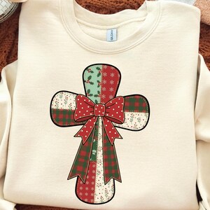 May include: Cream-colored sweatshirt featuring a patchwork cross design with a red polka-dot bow. The cross incorporates green holly, red plaid, and floral patterns. A festive and decorative apparel item.