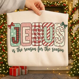 Könnte beinhalten: Cremefarbenes Sweatshirt mit dem Wort "JESUS" in bunten, festlichen Buchstaben. Die Buchstaben sind mit Karomustern, Streifen und Schneeflockenmustern verziert. Der Satz "is the reason for the season" ist darunter gedruckt.