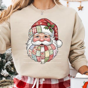 May include: Beige sweatshirt featuring a Santa Claus face design. The Santa face has a red and green plaid hat, a white beard, and a patchwork beard with floral and plaid patterns. A festive holiday-themed design.