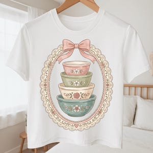 May include: White crop top featuring a pastel illustration of four stacked mixing bowls. The bowls are in shades of pink, green, and blue, decorated with floral patterns, and framed by a pink bow and a decorative oval border.
