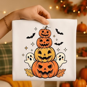 Może przedstawiać: Biały kwadratowy wzór na Halloween z stosu pomarańczowych dyni, nietoperzy, gwiazd i duchów. Dynie mają wyrzeźbione twarze. Jesienne liście u podstawy stosu. Wzór jest trzymany przez dłoń.