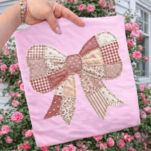 May include: A pale pink t-shirt with a large patchwork bow applique. The bow is crafted from various fabrics in pink, cream, and red hues, incorporating polka dots, gingham, and floral designs. The t-shirt is held up in front of a backdrop of pink roses.
