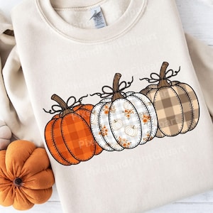 May include: A cream-colored sweatshirt featuring three decorative pumpkins. The pumpkins have different patterns: orange plaid, white floral, and brown plaid. Each pumpkin has a brown stem and a black bow.