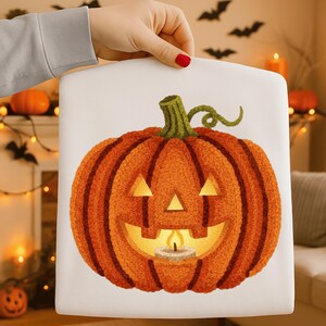 May include: A square, white Halloween-themed pillow featuring a carved orange pumpkin with a candle inside. The pumpkin has a green stem and a smiling face with triangular eyes. The pillow is held by a person.