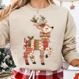 May include: Beige sweatshirt featuring a festive reindeer design. The reindeer is adorned with a patchwork pattern in red, green, and white, with holly, bows, and floral accents. The reindeer has a large bow around its neck and antlers decorated with leaves and flowers.