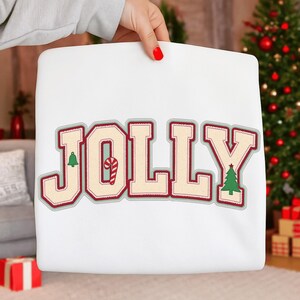 May include: A white cushion featuring the word "JOLLY" in large, festive lettering. The letters are cream-coloured with a red outline and a green border. The "O" features a candy cane, and the "L" has a Christmas tree.