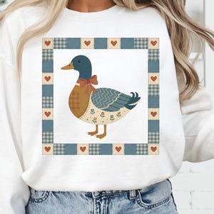 以下が含まれることがあります： 白いスウェットシャツ、漫画のアヒルのデザイン。アヒルは青い頭、茶色の体、赤いリボンをしています。デザインは、赤いハートが付いた青と白の格子縞のボーダーで囲まれています。スウェットシャツは柔らかい素材で作られています。