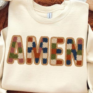 May include: Cream-colored sweatshirt with the word "AMEN" embroidered in large, colorful, patchwork-style letters. The letters are outlined in a tan color, and the patches include shades of blue, green, brown, and pink.