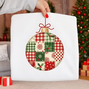 Może przedstawiać: Biała kwadratowa poduszka z dużym wzorem bombki choinkowej. Ozdoba to patchwork z czerwonych, zielonych i kremowych kwadratów z świątecznymi wzorami. Czerwona kokarda i złota nasadka dopełniają ozdobę.