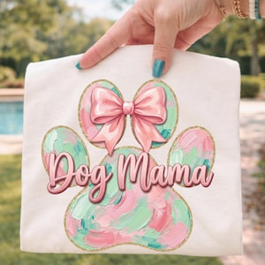May include: A white t-shirt featuring a colorful paw print design with a pink bow and the words "Dog Mama". The paw print has pink and green brushstrokes with gold glitter accents.