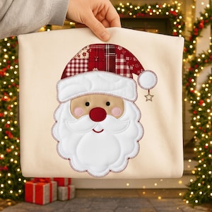 May include: A cream-colored Christmas pillow featuring a Santa Claus face. The Santa has a red nose, rosy cheeks, and a white beard and hat. The hat is made of a patchwork of red and white plaid fabrics. A gold star hangs from the hat.