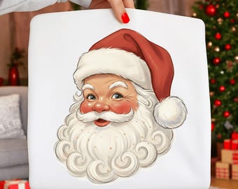 Op de afbeelding: Een wit vierkant kussen met een gedetailleerde illustratie van het gezicht van de kerstman. De kerstman wordt afgebeeld met een rode muts, roze wangen, een witte baard en een vrolijke uitdrukking. Het kussen wordt door een persoon vastgehouden.