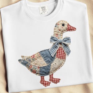 May include: White sweatshirt featuring a patchwork goose design. The goose has a floral head, blue gingham bow, and a body made of various patterned fabrics in red, blue, and cream. The goose's feet are red and white checkered.