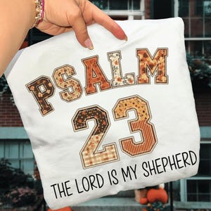 May include: White t-shirt with the text "PSALM 23 THE LORD IS MY SHEPHERD". The text is in a mix of fonts and patterns, including floral and plaid designs, in shades of brown and orange.