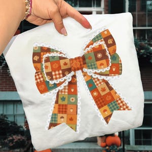 Può includere: Cuscino bianco con un grande design a fiocco colorato. Il fiocco presenta un motivo patchwork con elementi a tema autunnale come zucche, foglie e ghiande. Il fiocco è rifinito con pizzo bianco. Il cuscino è tenuto da una persona.