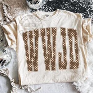May include: A cream-colored short-sleeve t-shirt featuring the letters WWJD in a large, brown, collegiate-style font with a white polka dot pattern. The letters are arranged in a slight arch across the center of the chest area of the garment.
