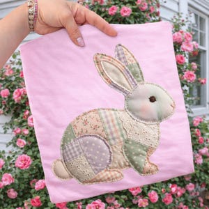 May include: A light pink fabric item featuring a patchwork bunny design. The bunny is made of various patterned fabrics in pastel colors, including floral, plaid, and polka dots. The background is a soft pink, and the item is held up in front of a rose bush.