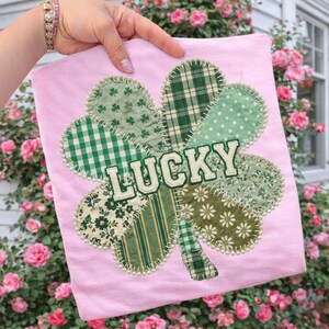 May include: A light pink t-shirt featuring a large, patchwork shamrock design. The shamrock is composed of various green patterned fabrics, including gingham, polka dots, and floral prints. The word "LUCKY" is stitched across the center in white letters.