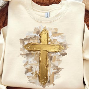 May include: Cream-colored sweatshirt featuring a gold cross design. The cross is painted with a textured, brushstroke effect, and is accented with gold flecks. The sweatshirt is a crew neck style.