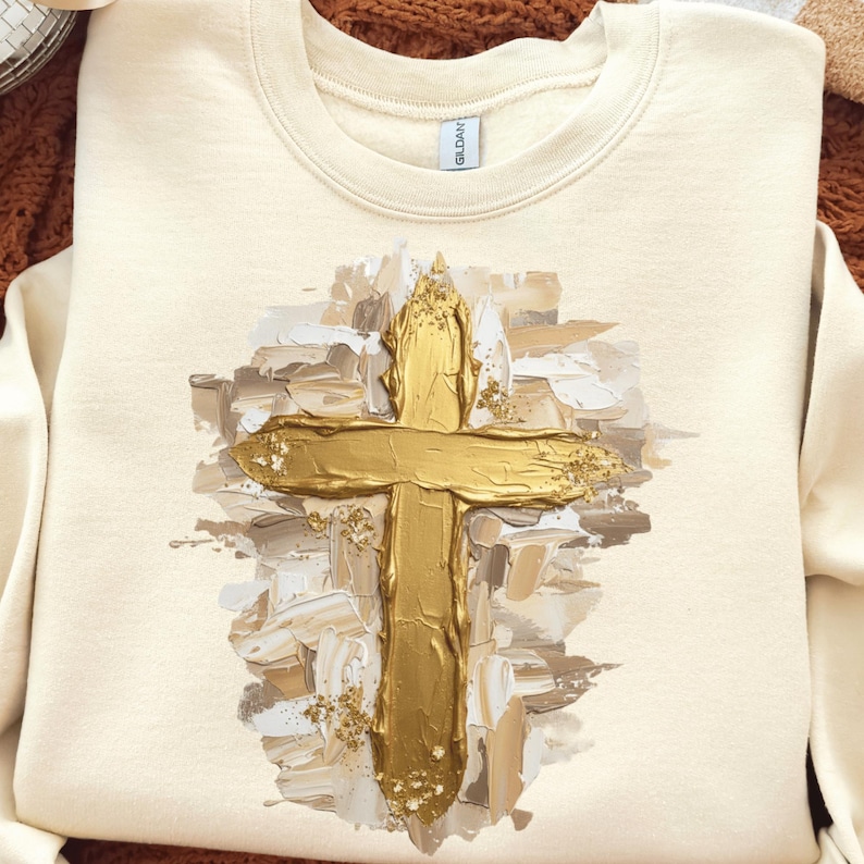 May include: Cream-colored sweatshirt featuring a gold cross design. The cross is painted with a textured, brushstroke effect, and is accented with gold flecks. The sweatshirt is a crew neck style.