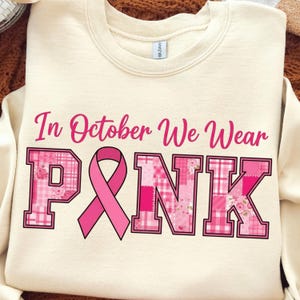 以下が含まれることがあります： クリーム色のスウェットシャツ。ピンクの筆記体で「In October We Wear PINK」と書かれています。「PINK」の文字はブロックレタースタイルで、中央にピンクのリボンがあります。文字は花柄とチェック柄で装飾されています。