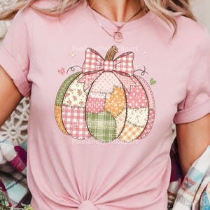 May include: Pink t-shirt featuring a patchwork pumpkin design. The pumpkin is composed of various patterned fabrics, including floral, gingham, and polka dots, with a large plaid bow. The design has a fall or autumn theme.