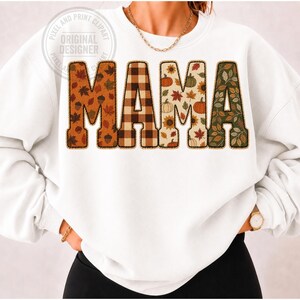 May include: A white crewneck sweatshirt featuring the word MAMA in large block letters across the chest. Each letter has a distinct autumn-themed pattern, including brown leaves, a brown and tan plaid, and various pumpkins and foliage.
