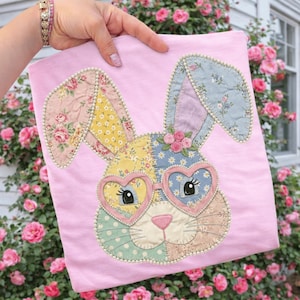 May include: A light pink t-shirt featuring a patchwork bunny face with heart-shaped glasses. The bunny has floral and polka dot patterned ears and face, with a pink nose and small rose accents. The design is embellished with a pearl-like trim.