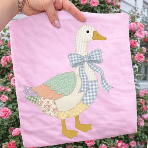 Könnte beinhalten: Rosa T-Shirt mit einem Patchwork-Gänsemotiv. Die Gans hat einen gestreiften Hals, eine blau-weiß karierte Schleife und bunte Patchwork-Flügel. Der Hintergrund ist ein zartes Rosa, und die Gans hat gelbe Füße.