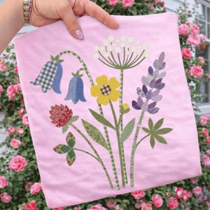 May include: A pink fabric item with an applique of various wildflowers. The design includes bluebell-shaped flowers, a yellow flower, and a purple lupine. Green leaves and stems complete the floral arrangement.