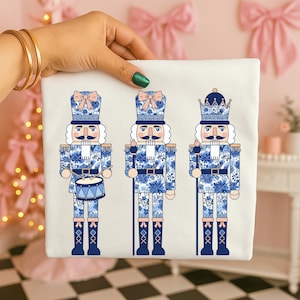 May include: A white square textile featuring three blue and white nutcracker soldier illustrations. Each soldier has a different hat, mustache, and uniform design. One soldier holds a drum, while the others hold a staff. The background is a pink color.