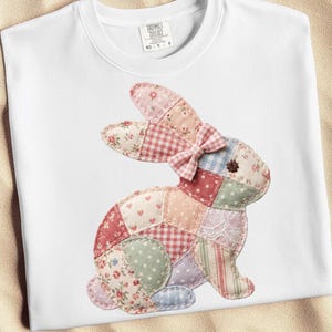 Puede incluir: Sudadera blanca con un diseño de conejo de patchwork. El conejo está hecho de varios patrones de tela, incluyendo floral, cuadros y lunares, con un lazo rosa. La sudadera tiene cuello redondo y mangas largas.